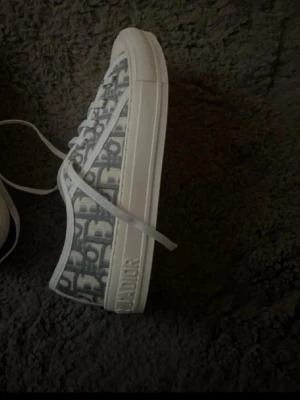 Dior gråvita låga sneakers - Snygga låga sneakers från Dior i grått och vitt med klassiskt Dior-mönster på canvas. Skorna har vita snören, gummisula och tydlig Dior-logga på sidan. Perfekta för dig som vill ha en lyxig och trendig look.