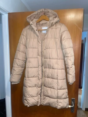Beige lång dunjacka från 157 - Säljer en beige lång dunjacka från 157 med huva och quiltad design. Jackan har dragkedja framtill och tryckknappar vid kragen. Perfekt för kalla vinterdagar och håller dig varm med sin vadderade konstruktion.