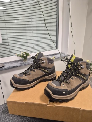 Bruna vandringskängor från Quechua 37 - Snygga bruna vandringskängor från Quechua med svarta och grå detaljer samt inslag av limegrönt. Kängorna har robust sula, metallhakar för snörning och är tillverkade i slitstarkt syntetmaterial och mocka. Perfekta för äventyr och outdoor.