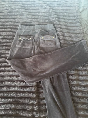 Mörkbruna mjukisbyxor i velour - Säljer ett par svarta mjukisbyxor i mjuk velour med raka ben. Byxorna har bred resår i midjan, två fickor fram och två bak med lock och dekorativa knappar. Perfekta för en chill och avslappnad stil.