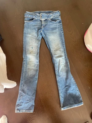 Blå bootcut jeans med bälte - Snygga blå bootcut jeans med slitningar och medföljande matchande jeansbälte. Klassisk femficksmodell och ljus tvätt för en avslappnad look. Perfekta till sneakers eller boots.