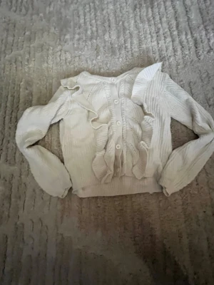 Vit kofta med volanger och knappar - Söt vit kofta med långa ärmar, knäppning framtill och dekorativa volanger längs knappraden. Stickad i mjukt material och har ribbade muddar vid ärmslut och nederkant. Perfekt för en trendig och feminin look.