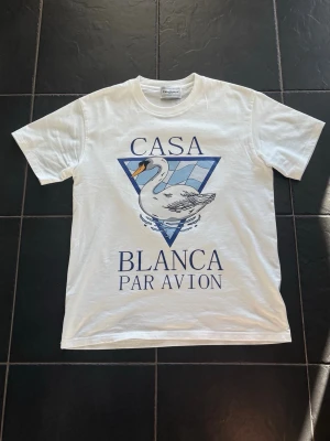 Vit Casa Blanca t-shirt med svan - Vit t-shirt från Casablanca med stort tryck av en svan och blå triangel på framsidan. Texten 'Casa Blanca Par Avion' i blått. Klassisk passform och rund hals. Perfekt för en stilren och trendig look.