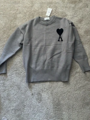 Grå stickad tröja från Ami Paris - Snygg grå stickad tröja från Ami Paris med svart hjärta och bokstaven A broderat på bröstet. Tröjan har rund halsringning och ribbade muddar vid ärmslut och nederkant. Perfekt för en stilren och trendig look.