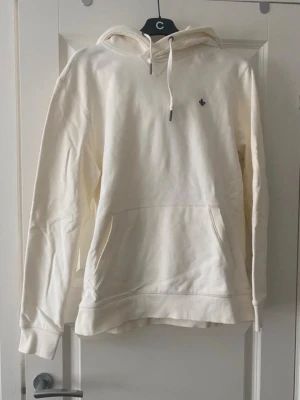 Vit Morris hoodie - Säljer en stilren vit Morris hoodie med huva och dragsnören. Hoodien har en stor magficka och ett litet diskret broderat märke på bröstet. Tillverkad i mjukt bomullstyg, perfekt för en avslappnad look. Passar dig som gillar enkel och clean stil.