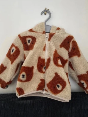 Teddyjacka med björntryck från Lilu - Mysig teddyjacka från Lilu i beige med brunt björnansikte-tryck. Jackan har huva, dragkedja framtill och är tillverkad i fluffigt teddy-material som håller dig varm. Perfekt för att sticka ut med ett gulligt och lekfullt mönster.