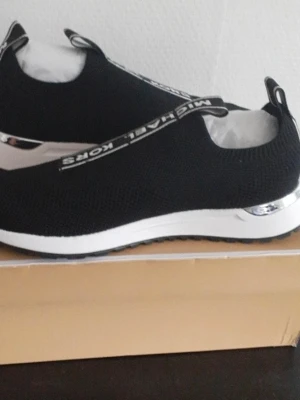 Svarta slip-on sneakers Michael Kors - Svarta slip-on sneakers från Michael Kors med vit och silverfärgad sula. Skorna har stickad mesh-överdel och tydliga logodetaljer på häl och dragflik. Perfekt för dig som gillar stilrena och sportiga skor med en touch av lyx.