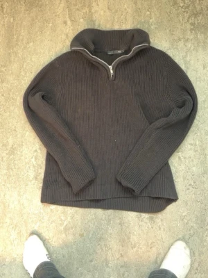Mörkgrå stickad tröja med halvzip - Mörkgrå stickad tröja från 157 med halvzip och hög krage. Tröjan har ribbstickad struktur, långa ärmar och en avslappnad passform. Perfekt för lager på lager och enkel att matcha med olika outfits.