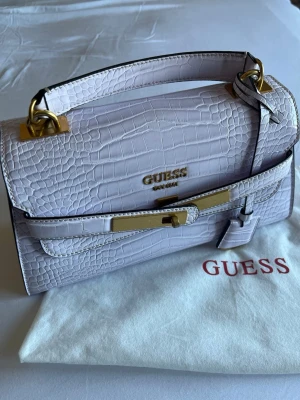 Ljuslila croco handväska från Guess - Snygg ljuslila handväska från Guess i croco-mönstrat konstläder med guldfärgade metalldetaljer. Väskan har ett topphandtag, dekorativt spänne framtill och Guess-logga i guld. Perfekt accessoar för att lyfta din outfit. Kommer med tillhörande axelrem, dustbag och originalbox. Lite tecken av användning på gulddetaljerna men det märks knappt. 