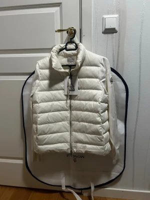Vit dunväst från Moncler - Snygg vit dunväst från Moncler med quiltad design och hög krage. Västen har dragkedja framtill och Moncler-logga på ärmen. Tillverkad i ett glansigt material med stickade detaljer på sidorna. Perfekt för lager-på-lager och stilren look.