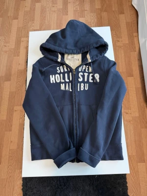 Hollister hoodie  - Mörkblå hoodie med dragkedja från Hollister. Stor vit text på bröstet med 'Surf Open Hollister Malibu'. Utrustad med huva och kängurufickor. Perfekt för en avslappnad och cool stil. Mjuk bomullsblandning för extra komfort. OBS armarna är inte vikt permanent!
