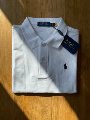 Vit pikéskjorta från Polo Ralph Lauren - Säljer en klassisk vit pikéskjorta från Polo Ralph Lauren med broderad logga på bröstet. Skjortan har krage, knappar framtill och är tillverkad i bomull. Perfekt för dig som gillar stilrena och tidlösa plagg.