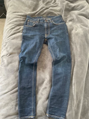 Blå smala jeans från Nudie Jeans - Säljer ett par klassiska blå jeans från Nudie Jeans med smal passform och snygga kontrastsömmar. Jeansen har fem fickor, normal midja och är tillverkade i slitstark denim. Perfekta för en avslappnad och trendig look.