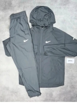 Grå Nike träningsset med huva - Snyggt grått träningsset från Nike med matchande byxor och jacka med huva. Setet har dragkedja framtill och vita Nike-loggor på både jackan och byxan. Tillverkat i lätt och funktionellt material, perfekt för träning eller chill.