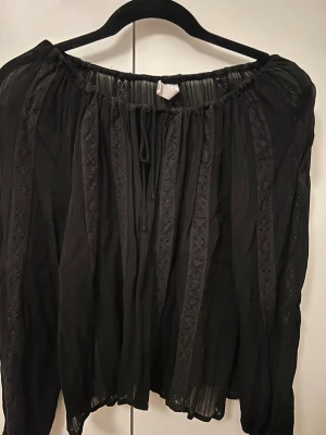 Svart boho-blus från Mango - Säljer en svart boho-inspirerad blus från Mango med transparent tyg och dekorativa spetsdetaljer i vertikala ränder. Blusen har resår vid halsen och långa, luftiga ärmar. Perfekt för dig som gillar en avslappnad men ändå trendig stil.