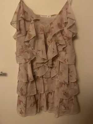 Ljus volangklänning från Stradivarius - Supersöt ljus kortklänning från Stradivarius med flera lager volanger och blommigt mönster i beige och rosa toner. Klänningen har tunna axelband och ett flowy fall, perfekt för sommarens alla tillfällen. Passar super bra vid axlarna och på kroppen.