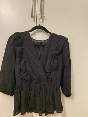 Svart blus med volanger från ONLY - Snygg svart blus från ONLY med djup V-ringning och volangdetaljer framtill. Blusen har trekvartsärm och är tillverkad i ett lätt, strukturerat tyg som ger en luftig känsla. Perfekt att styla med jeans eller kjol för en trendig look.