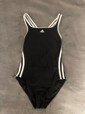 Svart baddräkt från Adidas - Sportig svart baddräkt från Adidas med vita ränder längs sidorna och vita kanter. Klassisk modell med öppen rygg. Tillverkad i snabbtorkande material, perfekt för simning, eller bara vanligt bad. Adidaslogga framtill, hör av er vid blir frågor!