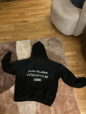 Svart hoodie Acne Studios Stockholm - Svart hoodie från Acne Studios med stor vit text på ryggen: 'Acne Studios STOCKHOLM 1996'. Tillverkad i mjuk bomull och polyesterblandning. Klassisk passform med huva och långa ärmar. Perfekt för en clean och stilren streetwear-look.