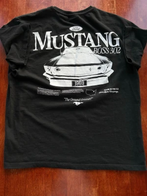 Svart Mustang Boss 302 t-shirt House - Svart t-shirt från House med Mustang Boss 302-tryck framtill. Klassisk rund hals och korta ärmar. Tillverkad i mjuk bomull och officiellt licensierad av Ford. Perfekt för dig som gillar bilar och vill ha en stilren look.