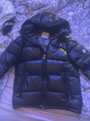 Svart dunjacka från Moncler med huva - Säljer en svart glansig dunjacka från Moncler med huva och dragkedja framtill. Jackan har två stora fickor med dragkedja och en patch på ärmen. Insidan har Monclers klassiska serietidningsmärke. Perfekt för kalla vinterdagar.