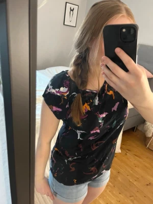 Svart topp med färgglatt mönster - Svart topp från Lindex med abstrakt färgglatt mönster i lila, orange, grön och vitt. Toppen har rund halsringning och korta ärmar. Perfekt till jeans eller shorts för en avslappnad look. Säljer en likadan men ljus topp (se sista bilden), har bundle rabatter!!!! 