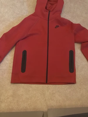 Röd Nike hoodie med dragkedja - Säljer en snygg röd hoodie från Nike med svart dragkedja och svarta detaljer på fickorna. Hoodien har huva och är tillverkad i mjukt material, perfekt för chill eller träning. Nike-loggan är tryckt på bröstet.