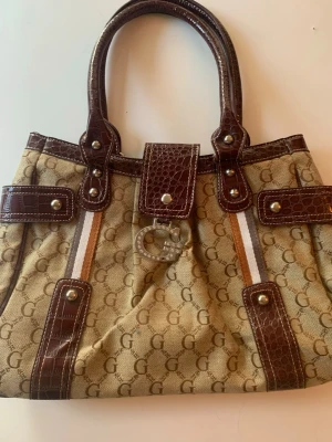 Beige och brun Guess märkt handväska - Väska med Guess märkning, täckt i G med mörk bruna detaljer i krokodilprint. Stort G spänne på framsida som saknar en sten men annars är i perfekt skik. Tre fack på insidan en med dragkedja. Väskan har två handtag och en drag kedja.