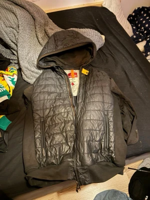 Svart vindjacka från Parajumpers - Snygg svart vindjacka från Parajumpers med quiltad front, huva och dragkedja. Jackan har två framfickor med dragkedja och ett coolt patch på ärmen. Perfekt för dig som vill ha en sportig och trendig look.
