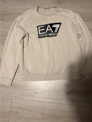 Beige sweatshirt från Emporio Armani - Säljer en beige sweatshirt från Emporio Armani EA7 med stor svartvit logga på bröstet. Tröjan har rund halsringning och långa ärmar. Perfekt för dig som gillar stilrena och trendiga plagg med tydlig logga.