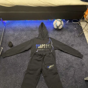 Svart hoodie och byxor från Trapstar - Säljer en svart hoodie och matchande byxor från Trapstar. Hoodien har en cool grafisk design med blå och vita detaljer på framsidan. Byxorna har samma design och en bekväm passform med dragsko i midjan. Perfekt för en avslappnad stil. Är upp till att sänka pris. Kan också göra byte!