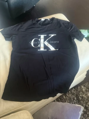 Svart Calvin Klein Jeans t-shirt - Snygg svart t-shirt från Calvin Klein Jeans med stort vitt CK-tryck på bröstet. Klassisk rund hals och korta ärmar, perfekt för en avslappnad och trendig look. Materialet är mjukt och känns skönt mot huden.