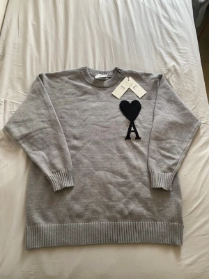 Grå stickad tröja från Ami Paris - Snygg grå stickad tröja från Ami Paris med svart hjärta och bokstaven A broderat på bröstet. Tröjan har rund halsringning, ribbade muddar och nederkant. Perfekt för dig som gillar stilrena och trendiga plagg.