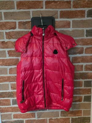 Röd dunväst med huva och logga - Snygg röd dunväst från Moncler med avtagbar huva och svart logotyp på bröstet. Västen har två dragkedjeförsedda fickor och quiltad design. Perfekt för lager-på-lager och håller dig varm under kyliga dagar.