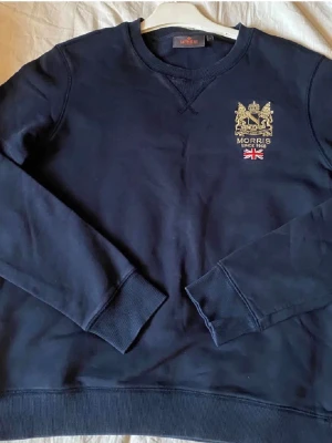  Morris - Mörkblå sweatshirt från Morris med guldbroderad logga och brittisk flagga på bröstet. Klassisk rund halsringning och ribbade muddar vid ärmslut och nederkant. Perfekt för en stilren och avslappnad look.