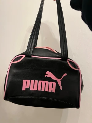 Svart och rosa Puma handväska   - Cool svart handväska med rosa detaljer och logga framtill. Väskan har dubbla handtag, dragkedja upptill och ljus insida. Tillverkad i konstläder med sportig look och retrovibbar. Perfekt för dig som gillar streetstyle och vill sticka ut. Inuti väskan ser den ganska använd ut och därför jag säljer den för billigt 