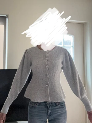 Grå ribbad kofta med puffärm - Snygg grå kofta med ribbad struktur och puffärmar. Koftan har rund halsringning och knäpps framtill med dekorativa knappar. Figurnära passform som markerar midjan och ger en feminin siluett. Perfekt att styla med jeans eller kjol.