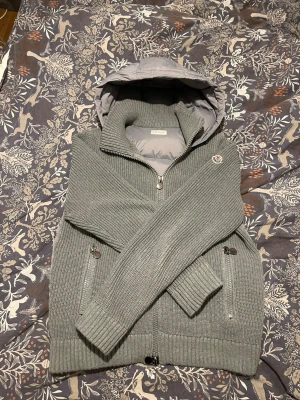 Grå stickad cardiganjacka Moncler - Säljer en grå stickad cardiganjacka från Moncler med vadderad huva och insida. Jackan har dragkedja framtill, ribbade muddar och två fickor med knappstängning. Klassisk Moncler-logga på ärmen och en riktigt snygg, clean look.