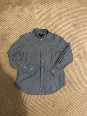 Blå skjorta från Polo Ralph Lauren - Snygg blå skjorta från Polo Ralph Lauren med klassisk button-down krage och broderad logga på bröstet. Skjortan har långa ärmar och knäppning framtill. Perfekt för en stilren look.