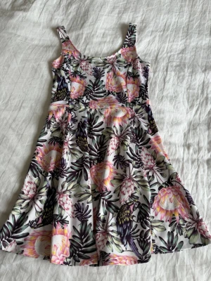 Blommig sommarklänning med axelband - Snygg kort klänning med blommigt mönster i rosa, grönt och svart på vit botten. Klänningen har smala axelband och en lätt utställd kjol, perfekt för varma dagar. Materialet känns mjukt och svalt, troligen bomull eller polyester.