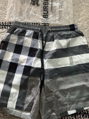Rutig badbyxa från Burberry - Snygga badbyxor från Burberry med klassiskt rutigt mönster i svart, vit och grå. Modellen har elastisk midja med snörning och en bakficka. Perfekt för strandhäng eller poolen, riktigt stilren look.