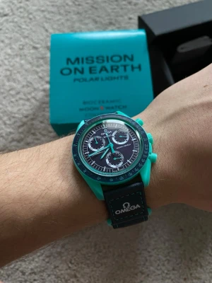 Omega x Swatch Mission on Earth Polar Lights klocka - Säljer en Omega x Swatch Mission on Earth Polar Lights klocka med turkos boett och svart armband. Klockan har kronograf och tydliga vita detaljer på urtavlan. Levereras i originalbox med tillbehör. Perfekt för dig som gillar unika accessoarer.