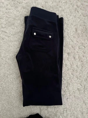 Mörkblå velourbyxor Juicy Couture - Mörkblå mjukisbyxor från Juicy Couture i mjuk velour. Byxorna har en bakficka med lock och silverfärgade knappar samt bred resår i midjan. Perfekta för en chill och avslappnad stil.