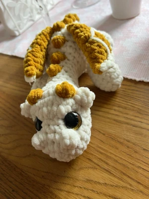 Vit drake med gulbruna detaljer - Denna urgulliga drake längtar efter sitt för alltid hem🤍🫶🏼✨ Det är en vit drake med gulbruna detaljer, virkat med mjukt garn från Hobbii som heter Baby Snuggle🫶🏼 Den har hårda guldiga ögon som inte går att ta av, sitter som bergen✨💛 Den är cirka 30 cm lång💛