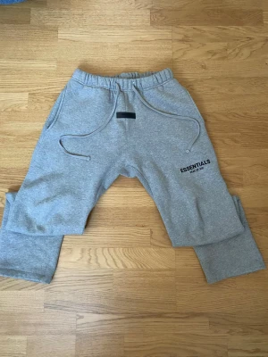 Grå Essentials Fear of God mjukisbyxor - Säljer ett par grå mjukisbyxor från Fear of God Essentials med dragsko i midjan och fickor på sidorna. Byxorna har en relaxed fit och logga tryckt på vänster lår. Perfekta för en chill och avslappnad stil.