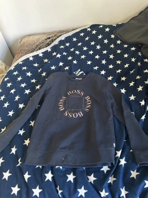 Mörkblå sweatshirt från Hugo Boss - Mörkblå sweatshirt från Hugo Boss med rund halsringning och tryckt BOSS-logga i cirkelform på bröstet. Tröjan har långa ärmar och är tillverkad i mjukt bomullsmaterial för en bekväm känsla. Perfekt för en avslappnad och stilren look.