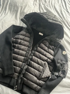Svart pufferjacka från Moncler - Snygg svart pufferjacka från Moncler med quiltad front och huva. Jackan har stickade ärmar och sidor, dragkedja framtill och en ficka med Moncler-logga på ärmen. Perfekt för kalla dagar och riktigt trendig stil.