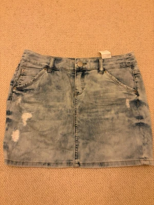 Ljusblå jeanskjol - Trendig ljusblå jeanskjol från S.oliver med slitna detaljer framtill. Kjolen har klassisk femficksdesign, bälteshällor och knappstängning. Perfekt för dig som gillar en avslappnad och cool stil.