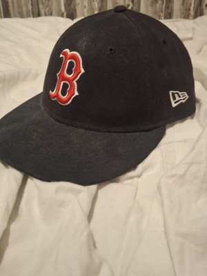 Boston Red Sox keps från New Era - Säljer en svart Boston Red Sox keps från New Era med klassisk böjd skärm och broderad röd B-logga framtill. Kepsen är tillverkad i bomull och har New Era-loggan broderad på sidan. Perfekt för dig som gillar sport och streetstyle.