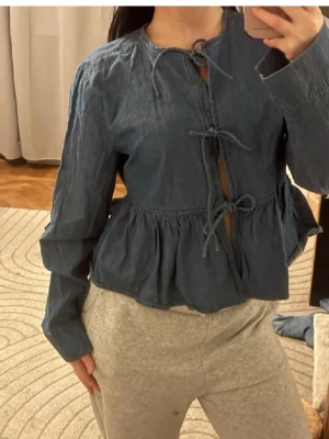 Superpopulära slutsålda jeanstoppen❤️ - Snygg blå blus i denimliknande material med volang nedtill och tre knytband framtill. Blusen har långa ärmar och rund halsringning. Perfekt för en trendig och avslappnad look. Köptes för 1000 kr. Passar även xs/s